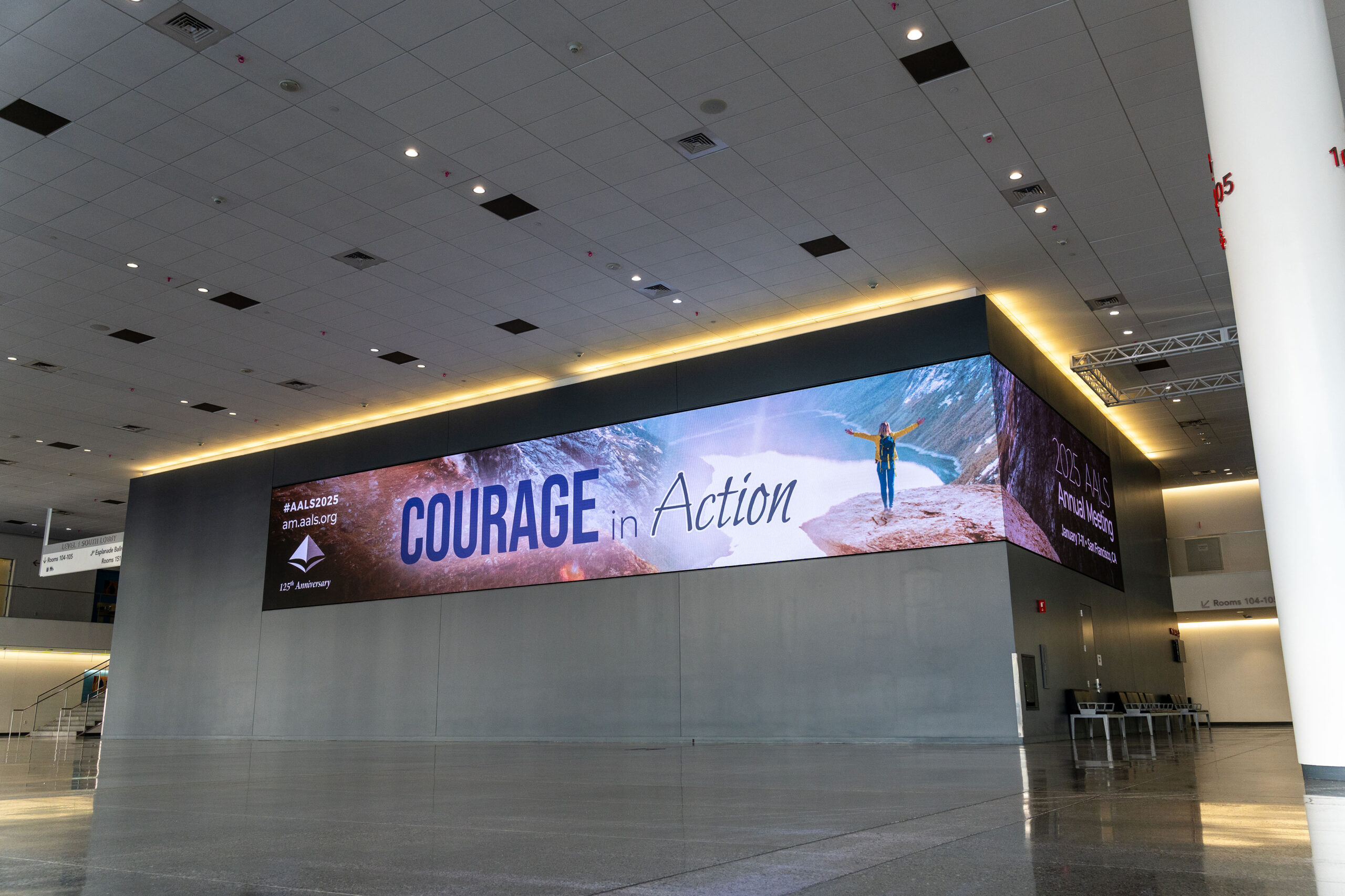 a large grey meeting space lobby with a purple and blue wrap banner reading "Courage in Action" with a white woman in a yellow sweater celebrates at the top of a summit